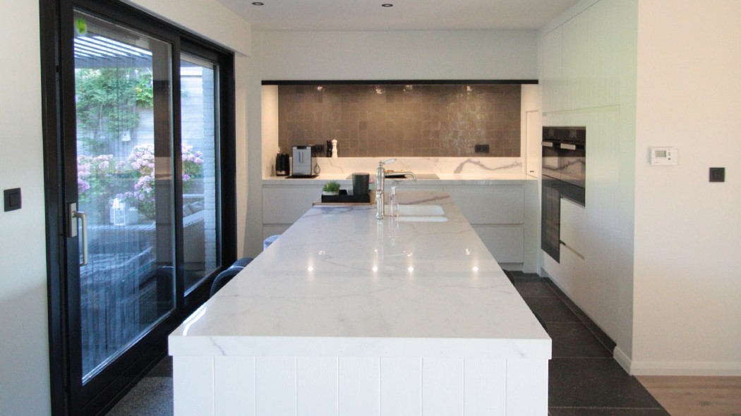 foto van een moderne keuken, wit keukeneiland in het midden met een marmeren werkblad. Achteraan de keuken nog extra werkruimte op op maat gemaakte kasten en langst de zijkant een schuifdeur richting terras