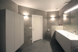 Totaal overzicht van een moderne badkamer. Grijze tegels op de vloer en tegen de muur. Tegen de linker muur hangt een wandkast, aan de rechterkant een lavabomeubel met 1 grote lavabo en 2 kranen. daarboven een spiegel met licht. De verlichting in de badkamer wordt verzorgt door lichtelementen tegen de muur