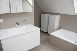 foto van een moderne badkamer onder de schuine kant van het dak. Ligbad, lavabo en kastjes. Op de vloer en tegen de muren werden keramische tegels gebruikt. Boven de lavabo, waaronder er twee schuiven in een hangkast hangen, hangt een spiegelkast.