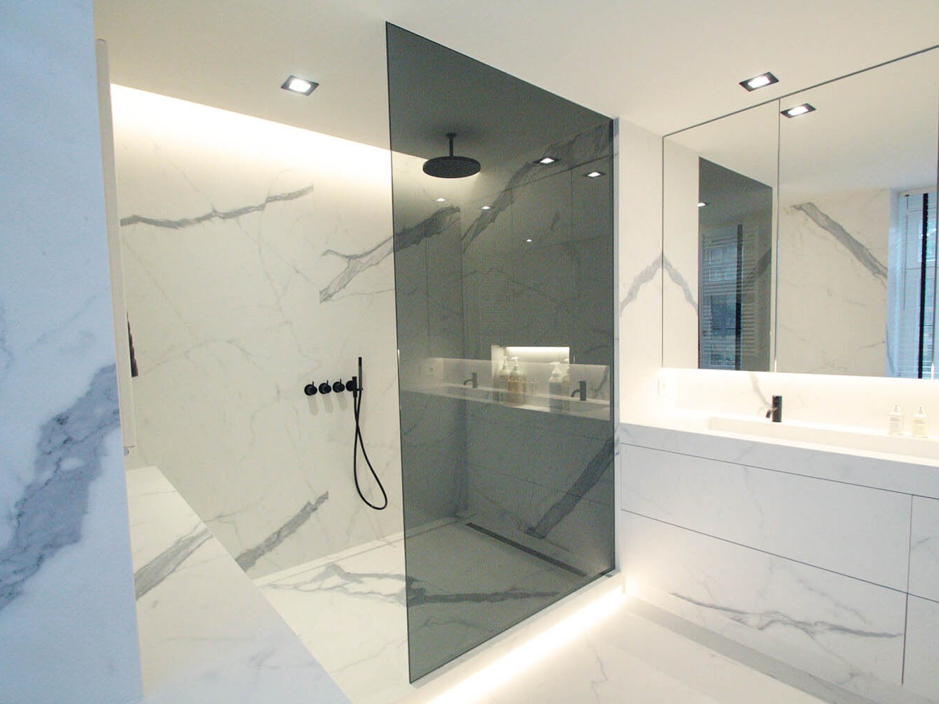 foto van moderne badkamer, foto van de hoek met douche en een deel van de wastafel met daarboven spiegelkasten. op de muur en grond werden keramische grootformaat tegels gebruikt met een marmer effect