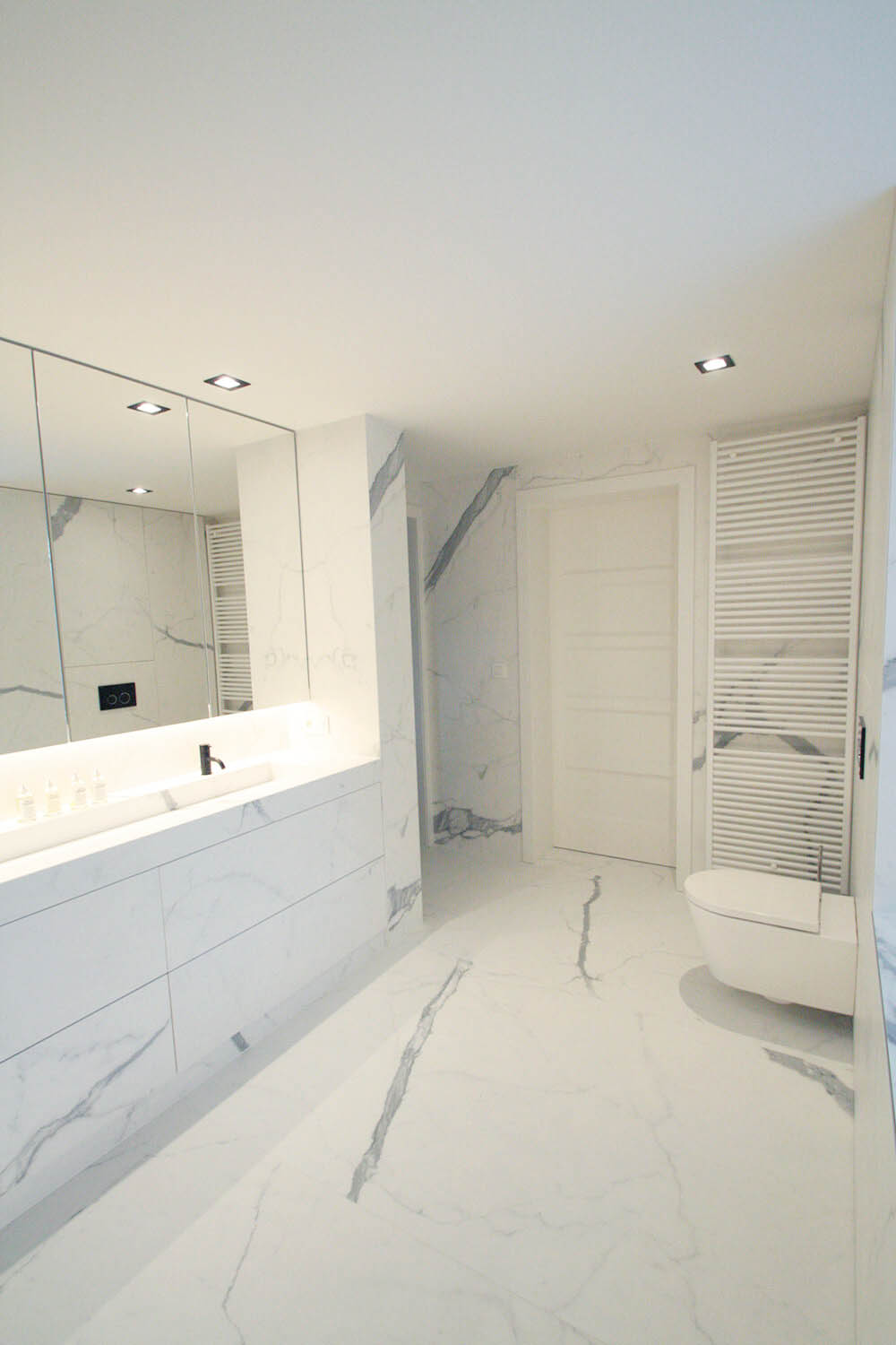foto van witte moderne badkamer met tegels op de vloer en tegen de muur, ook de kasten werden met dit materiaal afgewerkt. Het gebruikte materiaal heeft een marmer-effect
