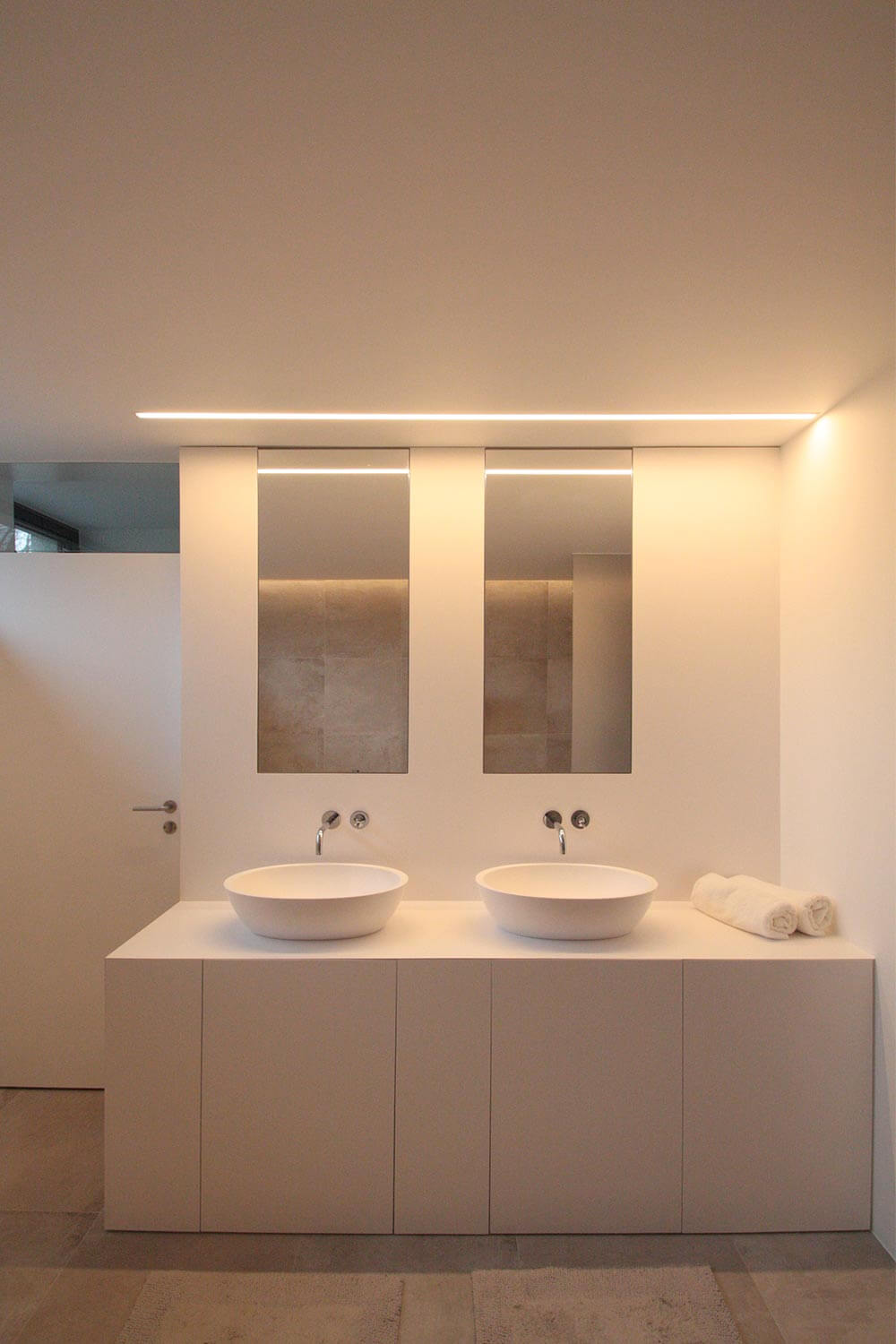 moderne badkamer met beige keramische tegels op de vloer. Witte muren. Zicht op muur met het lavabomeubel dat bestaat uit een kast met deuren. De lavabo's op de kast zijn vrijstaand. boven de lavabo's hangen spiegels