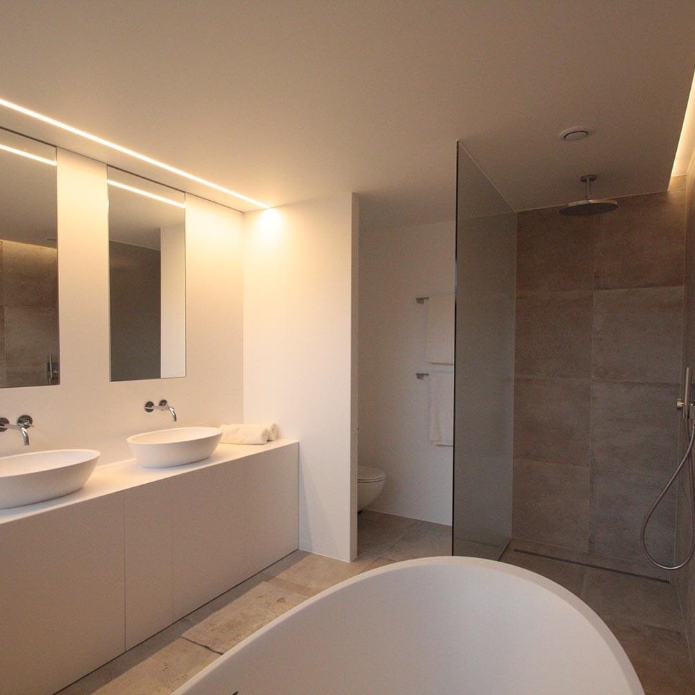 foto van een moderne badkamer getrokken van achter het vrijstaande bad. Recht vooruit rechts zie je de inloopdouche met regendouchekop, links een toilet achter een muur, aan de muur handdoekrekken en rechts lavabomeubel met spiegels boven
