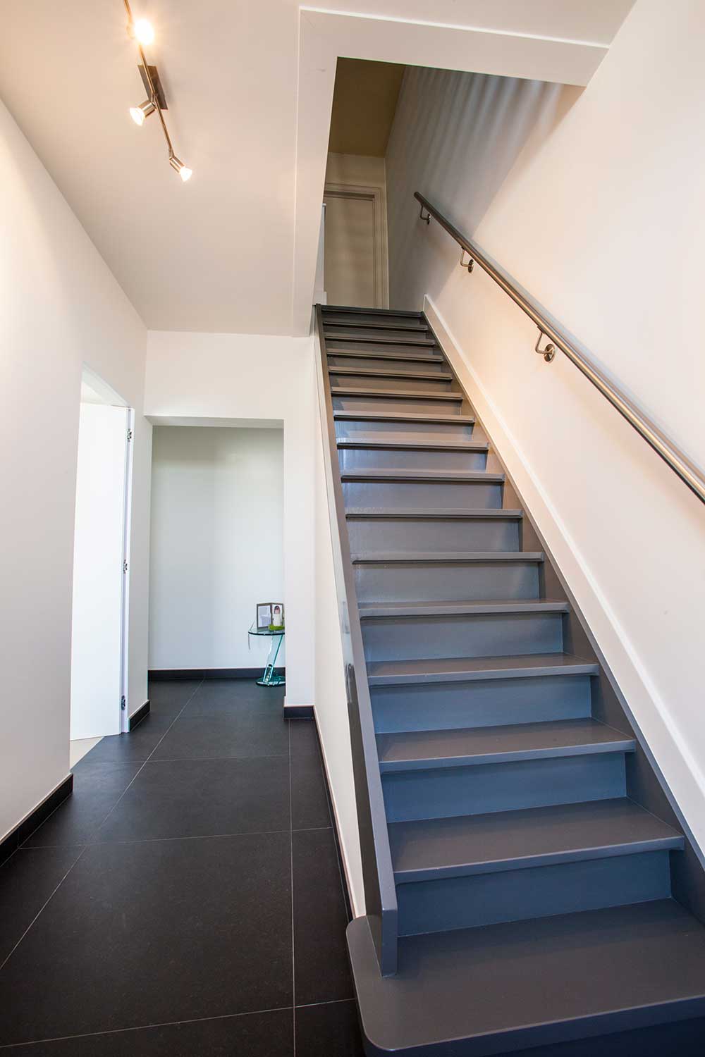 foto van een moderne hal. Grijs geverfde trap, witte muren en donker grijze tegels op de vloer. de trapleuning tegen de muur is in aluminium