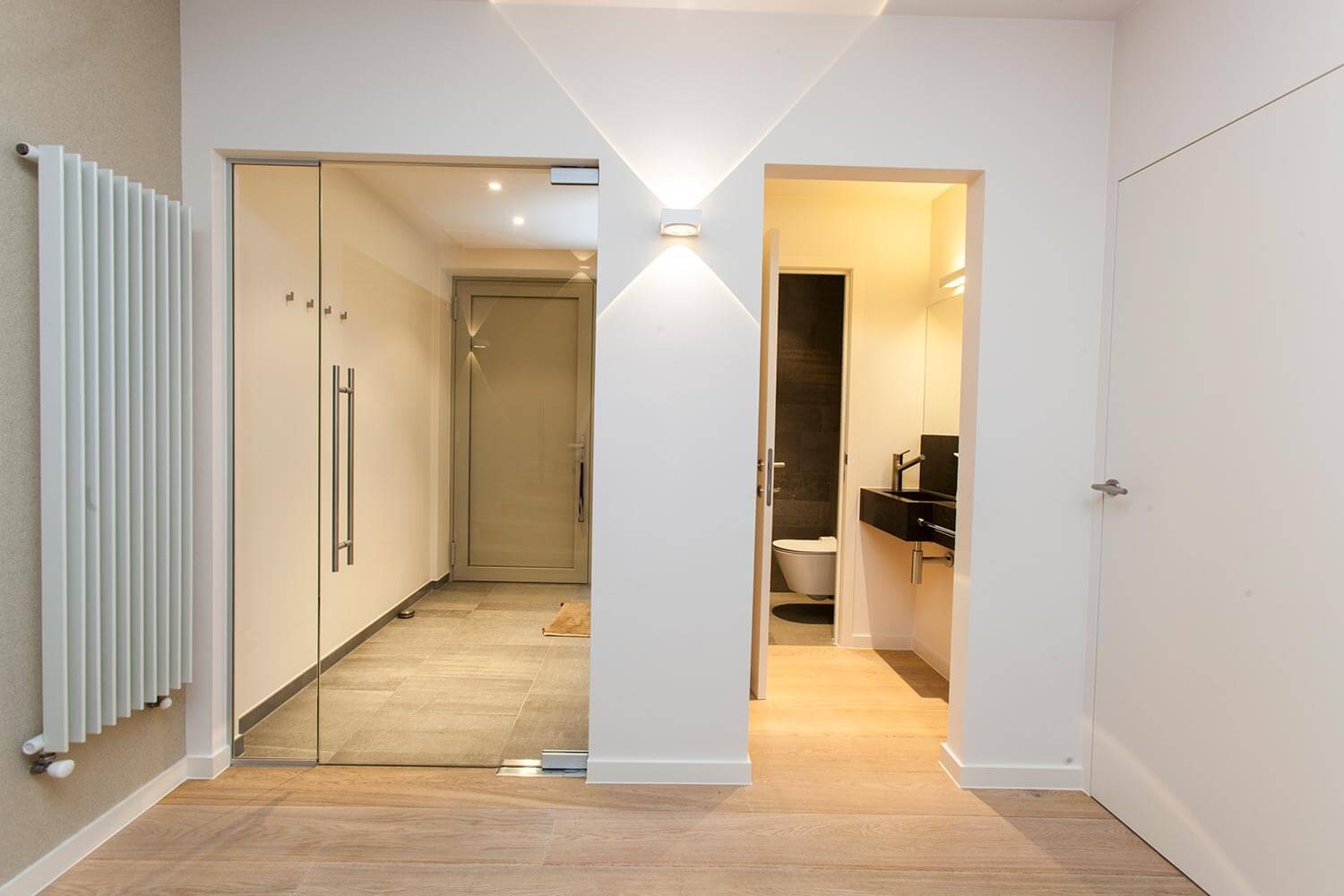 foto van een moderne witte inkomhal, met legklaar parket op de vloer, glazen deur richting de voordeur en doorgang richting een gastentoilet. wandverlichting en een moderne centrale verwarming tegen de muur